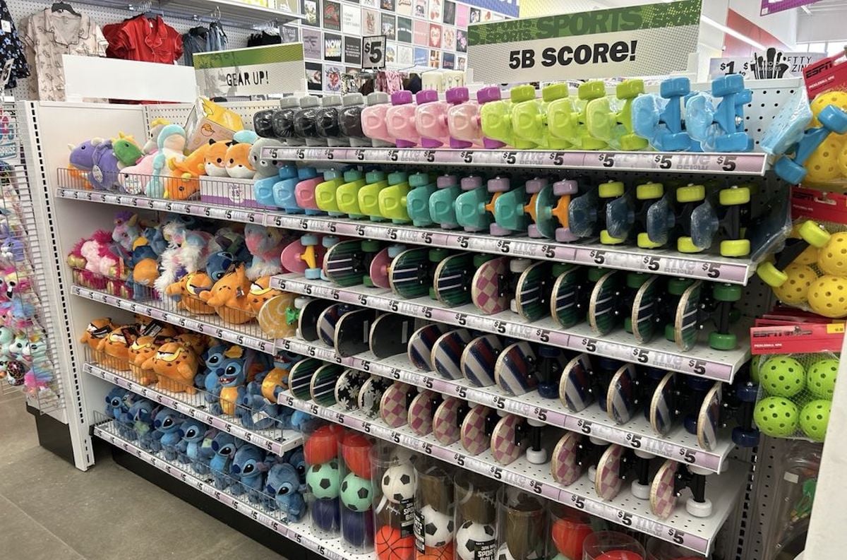Shelves in a store display colorful piggy banks, plush toys, and sports-themed water bottles shaped like soccer balls, basketballs, and footballs. For anyone’s first time at Five Below, overhead signs read "5B SCORE!" as the floor gleams below.