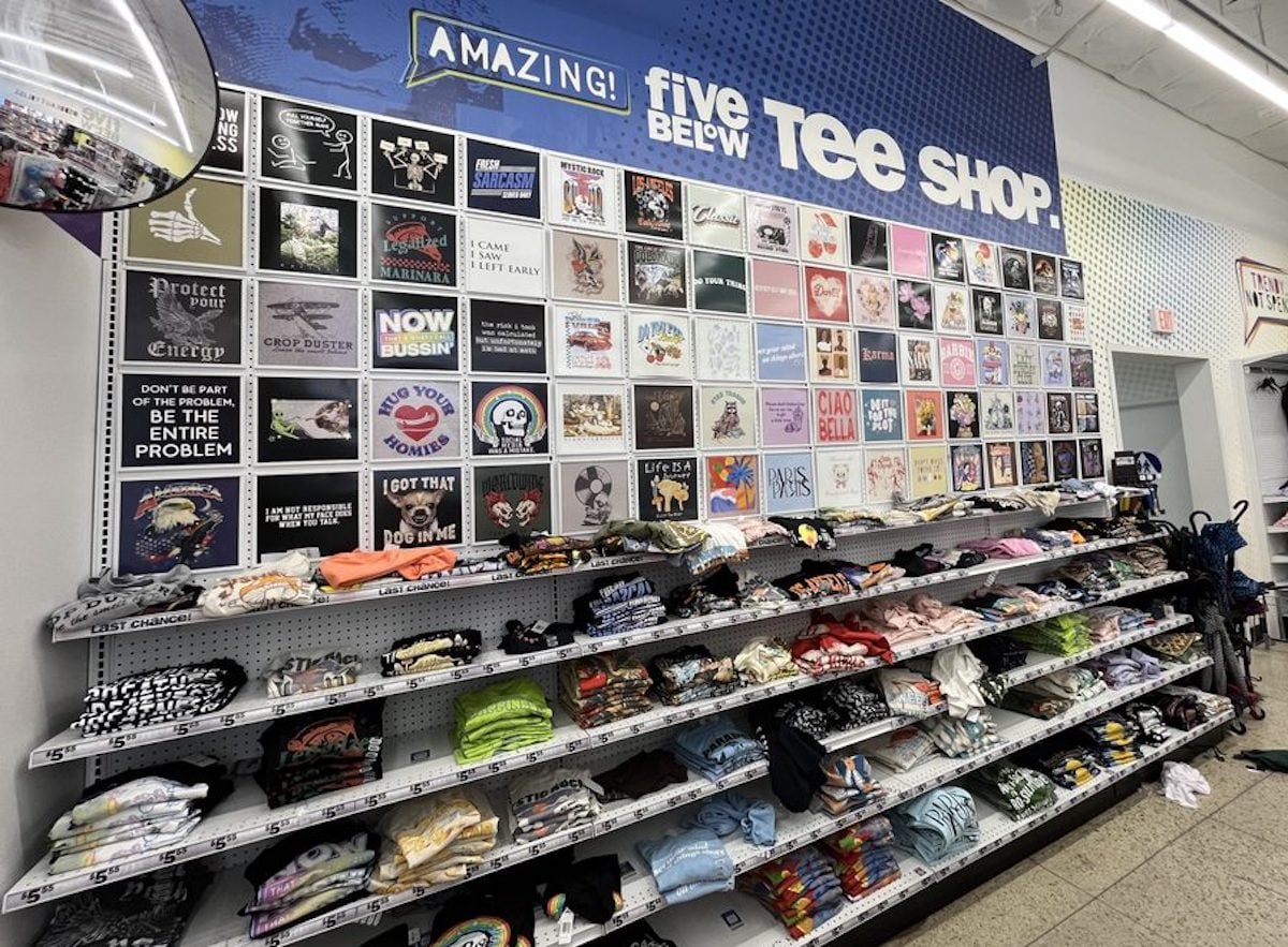 A display wall at Five Below's Tee Shop welcomes those visiting for the first time at Five Below, showcasing graphic T-shirt designs above shelves of folded tees. Signs and mirrors complete the look in this brightly lit store.