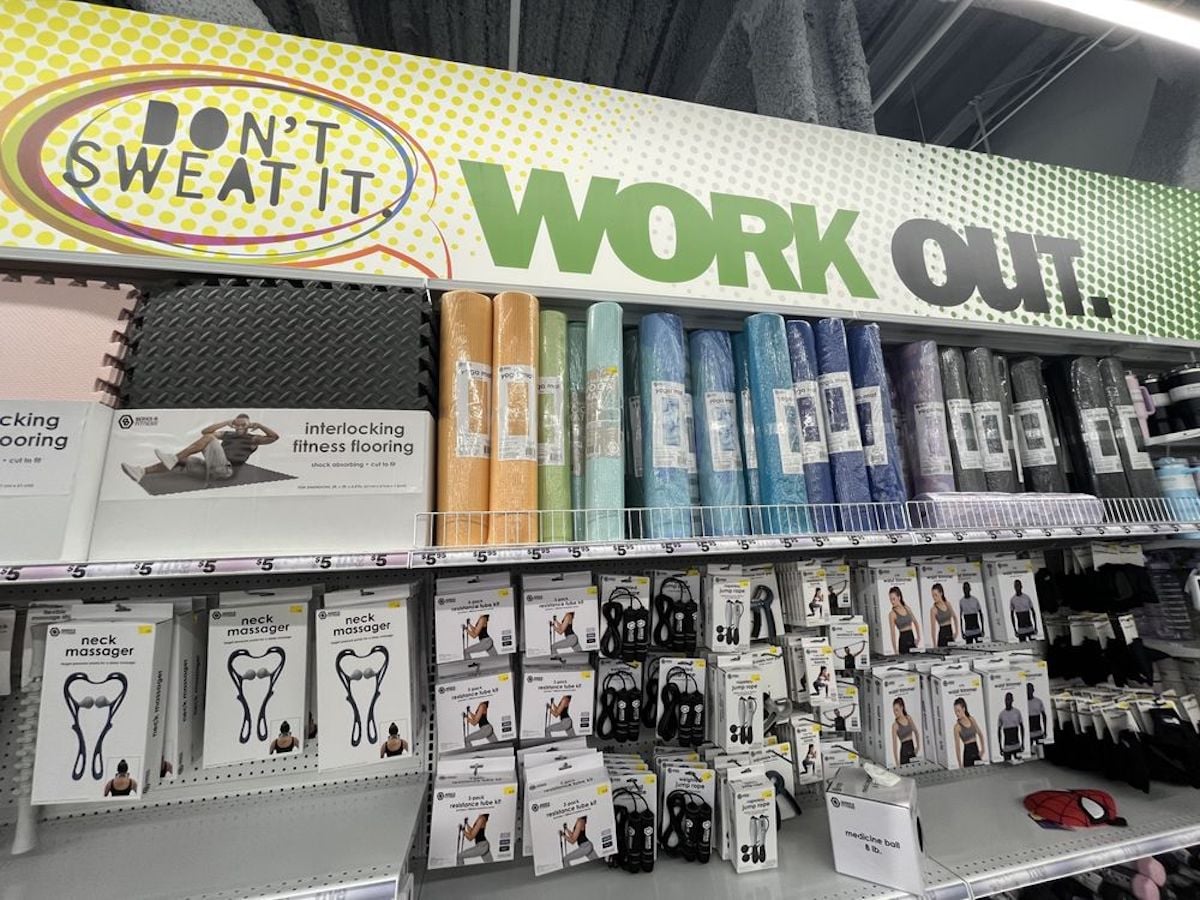 Shelves display foam exercise mats in various colors and packaged fitness accessories. Above, a sign reads “DON’T SWEAT IT” and “WORK OUT.” The store’s bright lighting and gray walls invite excitement for your first time at Five Below.