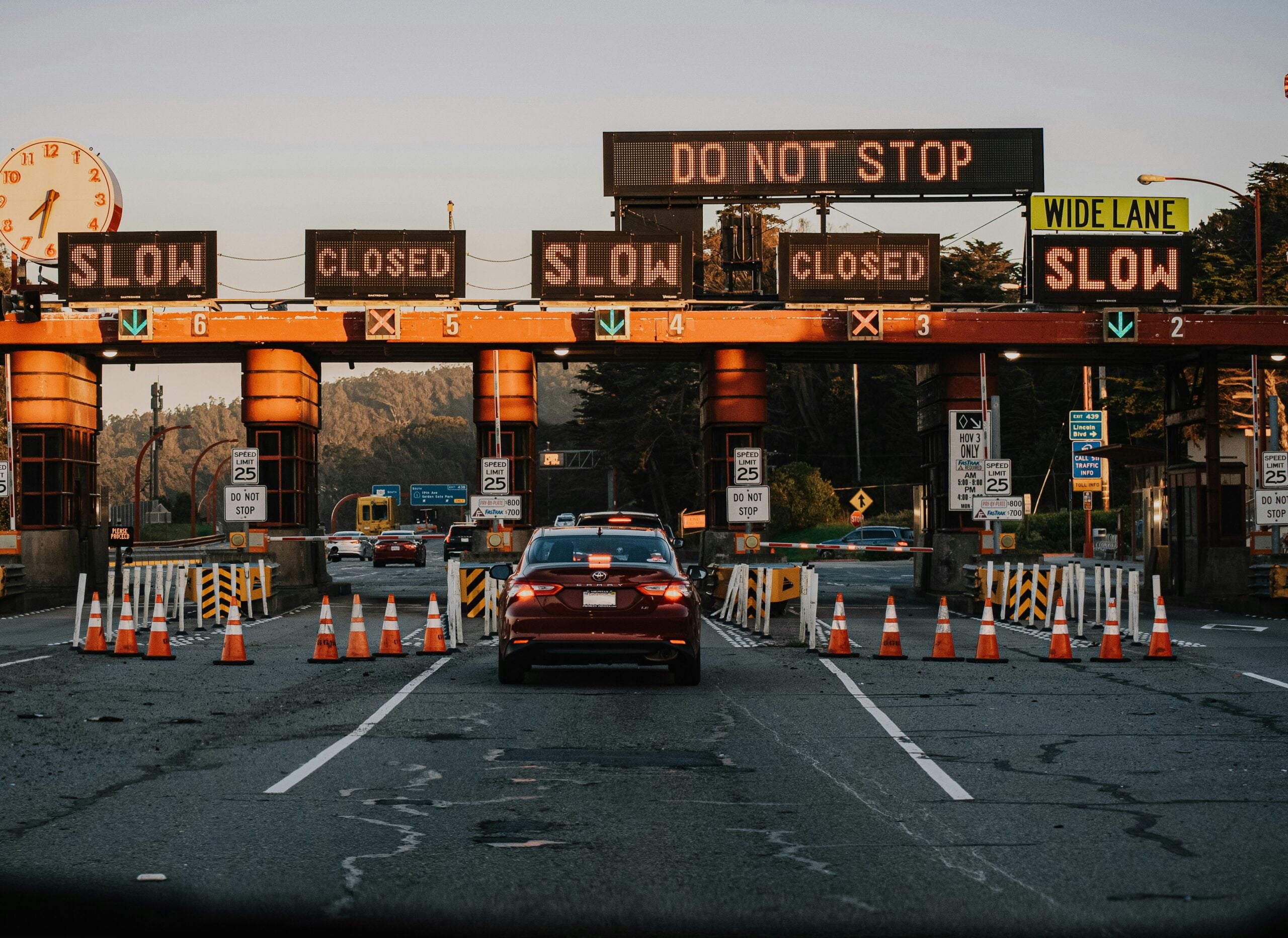 Cars approach a toll booth with multiple "CLOSED" and "SLOW" signs. Orange traffic cones guide vehicles, and an electronic sign above reads "DO NOT STOP." Speed limit signs indicate 25 mph. Trees are visible in the background.