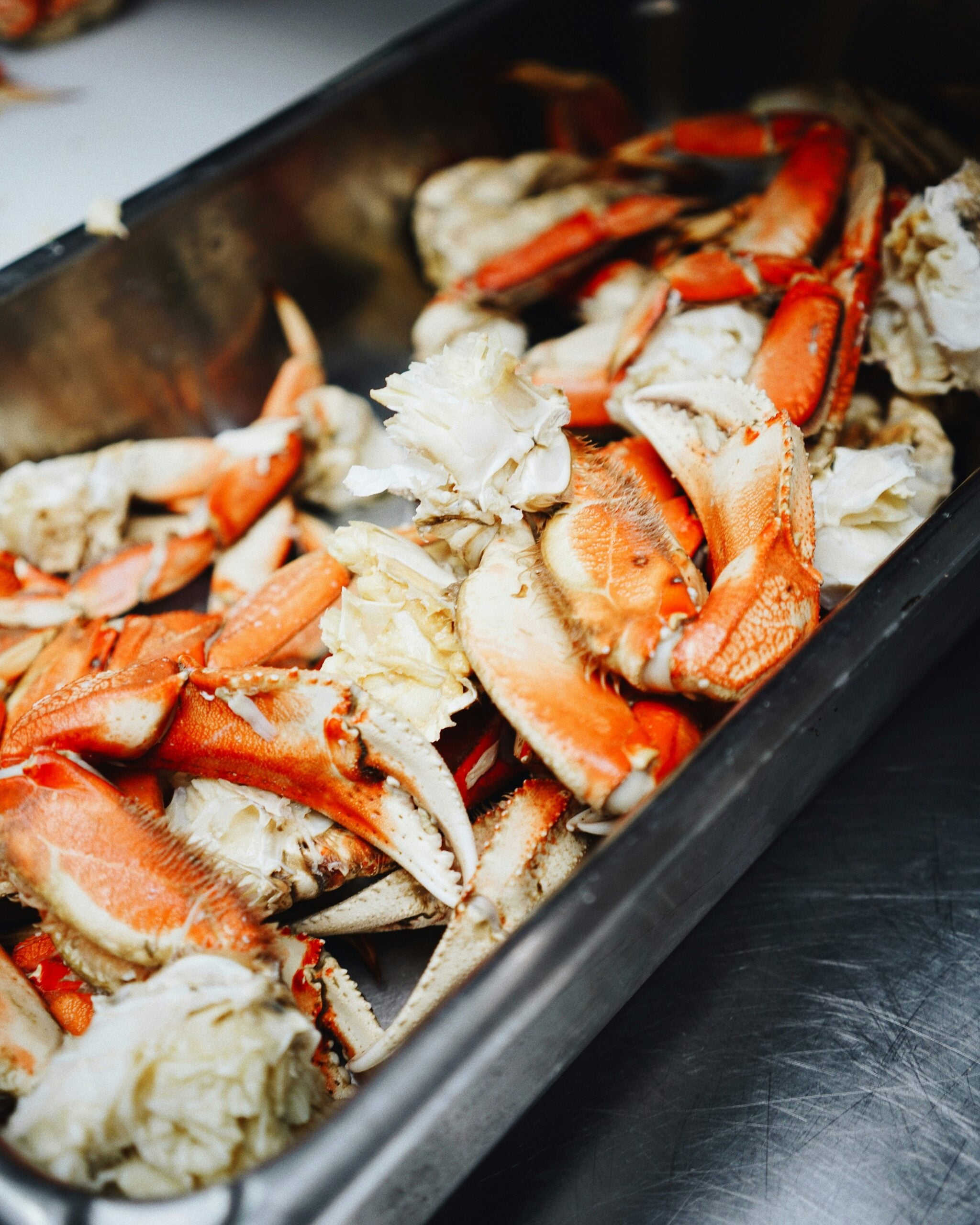 A metal tray filled with freshly cooked crab legs and claws, displaying vibrant orange shells and white meat, placed on a stainless steel countertop.