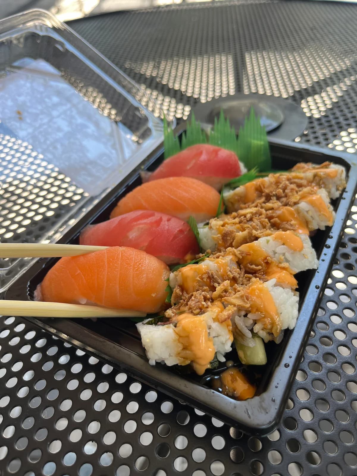 A black plastic takeout container holds a variety of sushi, including nigiri with salmon and tuna, and a roll topped with crispy onions and a creamy sauce. Chopsticks are picking up a piece of salmon nigiri.