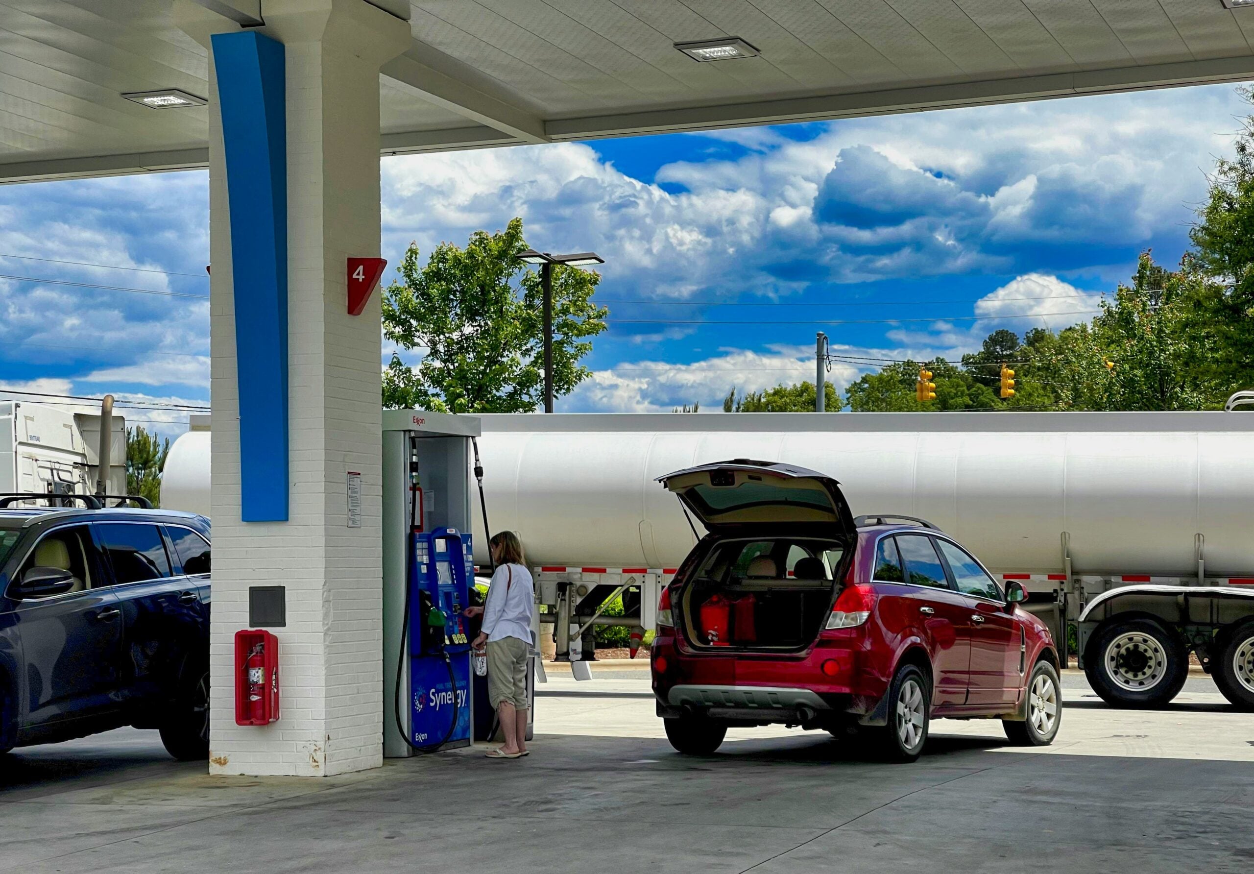 A woman stands at a gas pump refueling a red SUV with its trunk open at a gas station. A blue car and a large white tanker truck are also visible under a cloudy sky. best day to buy gas.