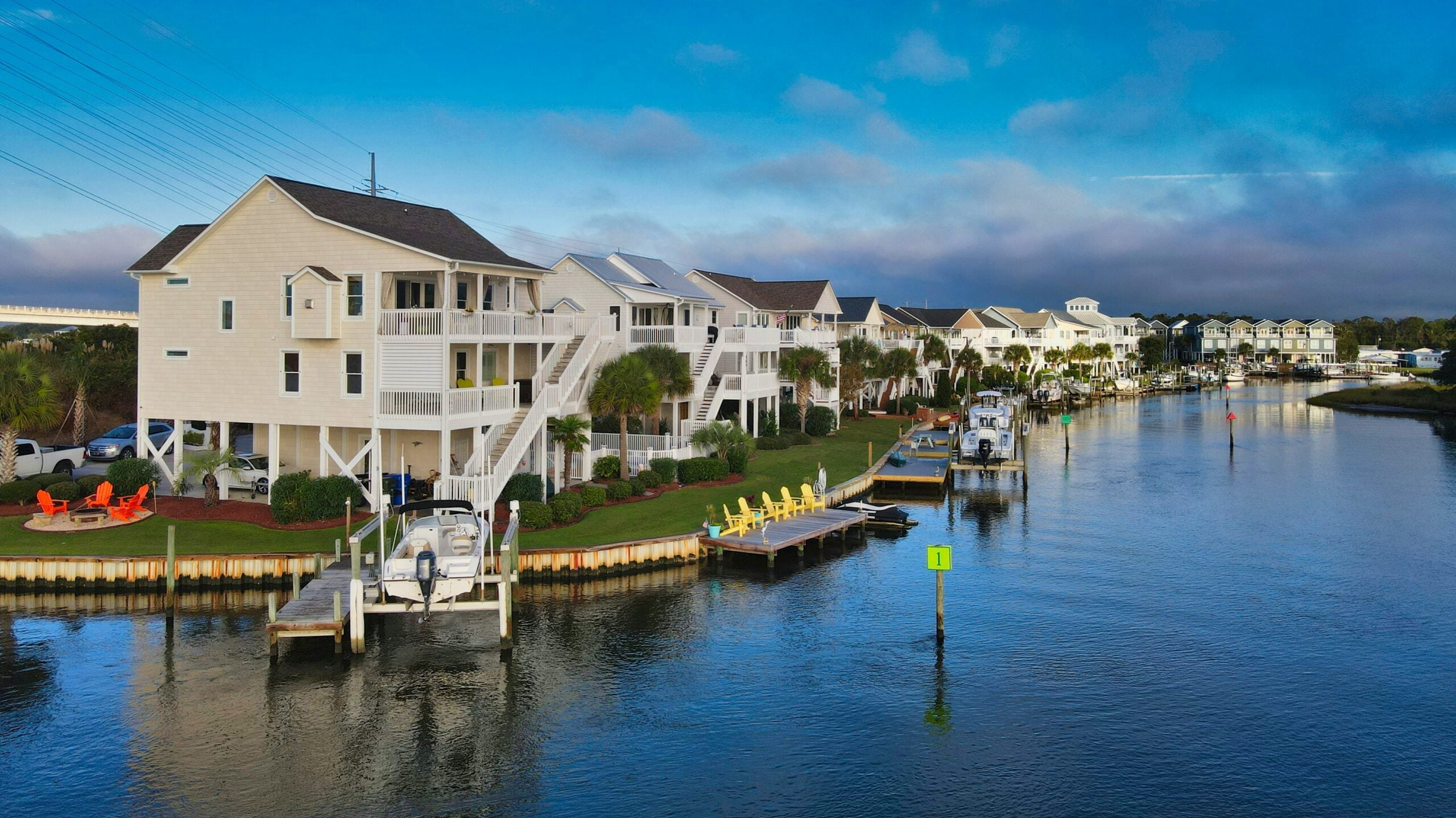Row of waterfront houses with docks, boats, and colorful outdoor chairs lining a calm canal under a partly cloudy blue sky. Lush green lawns and palm trees complete the peaceful neighborhood scene.