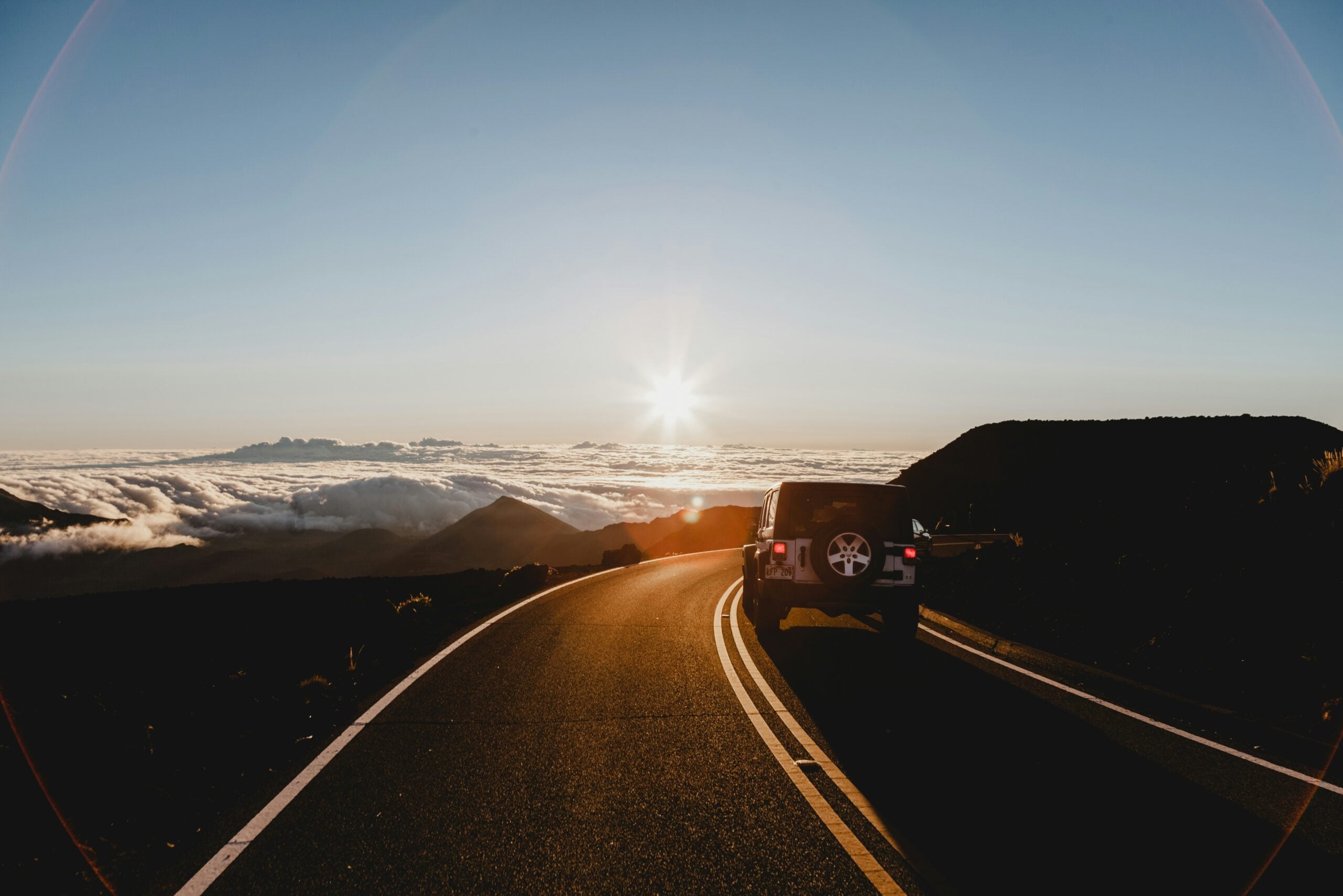 A car parked on the side of a winding mountain road at sunset, overlooking clouds and distant peaks under a clear sky. The sun is setting on the horizon, casting a warm glow.