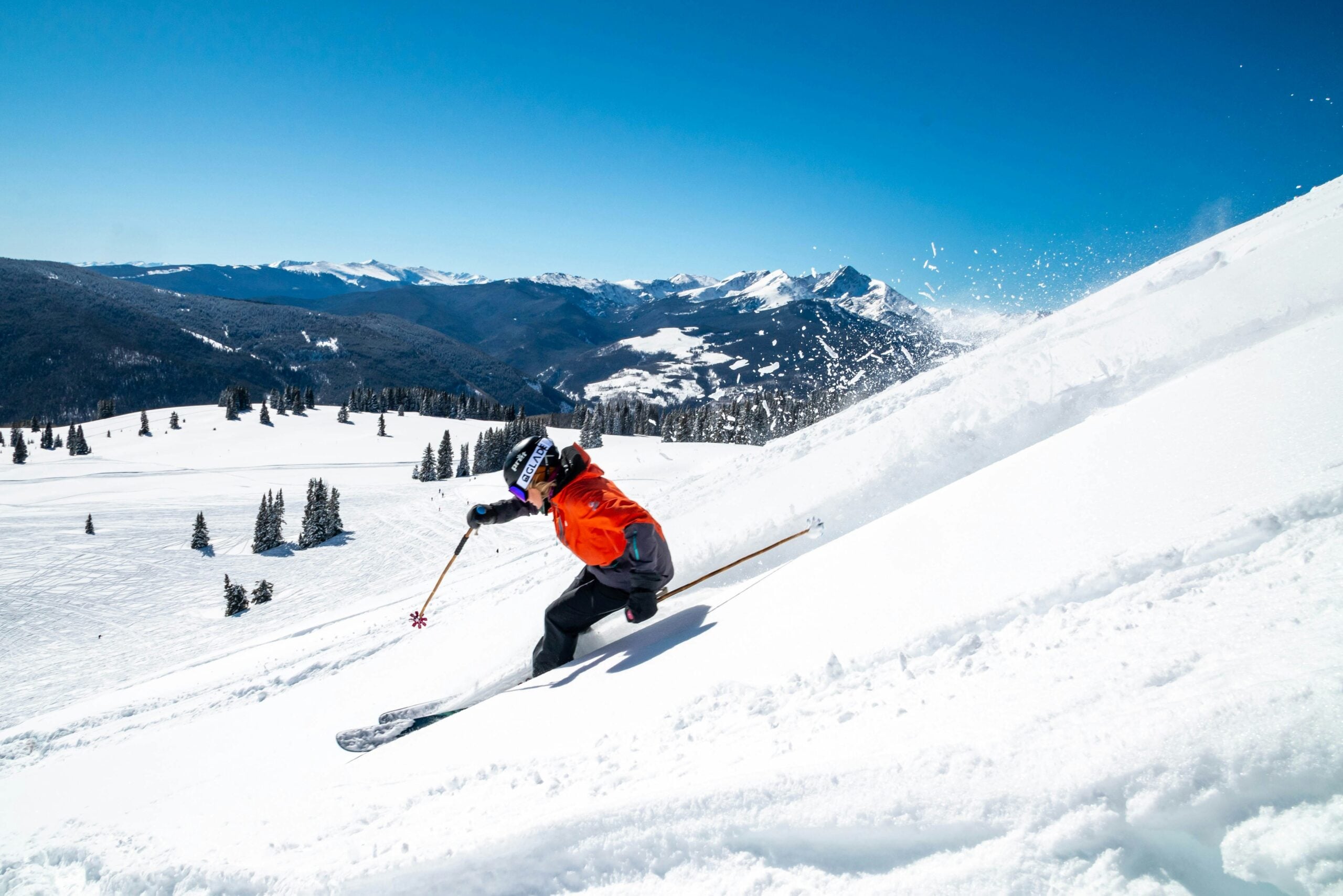 A skier in a red jacket and helmet carves down a snowy mountain slope on a sunny day, with snow spraying behind and forested mountains visible in the background under a clear blue sky.