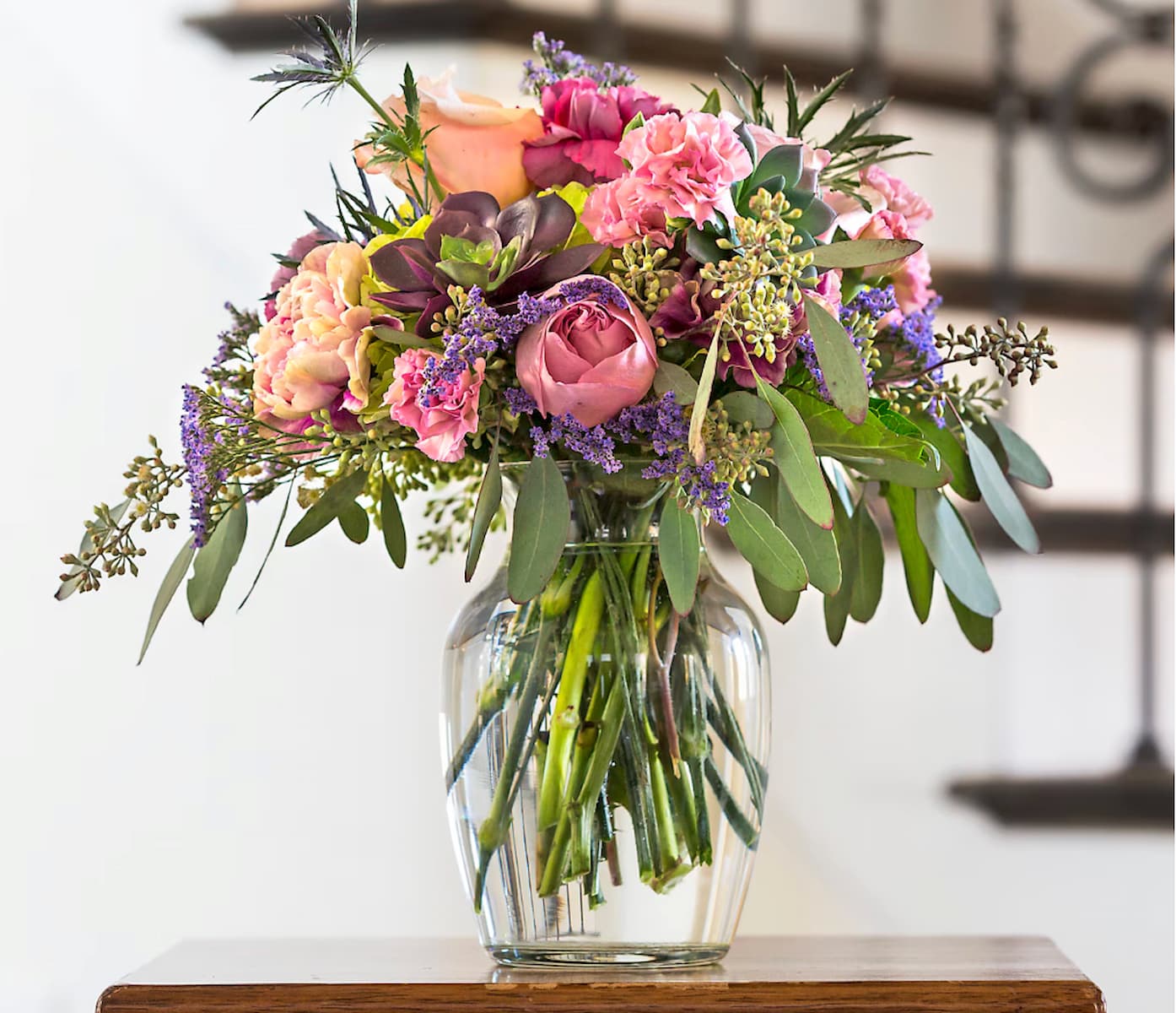 A glass vase filled with a bouquet of pink, purple, and peach flowers, accented with green foliage, sits on a wooden surface with a blurred staircase in the background.