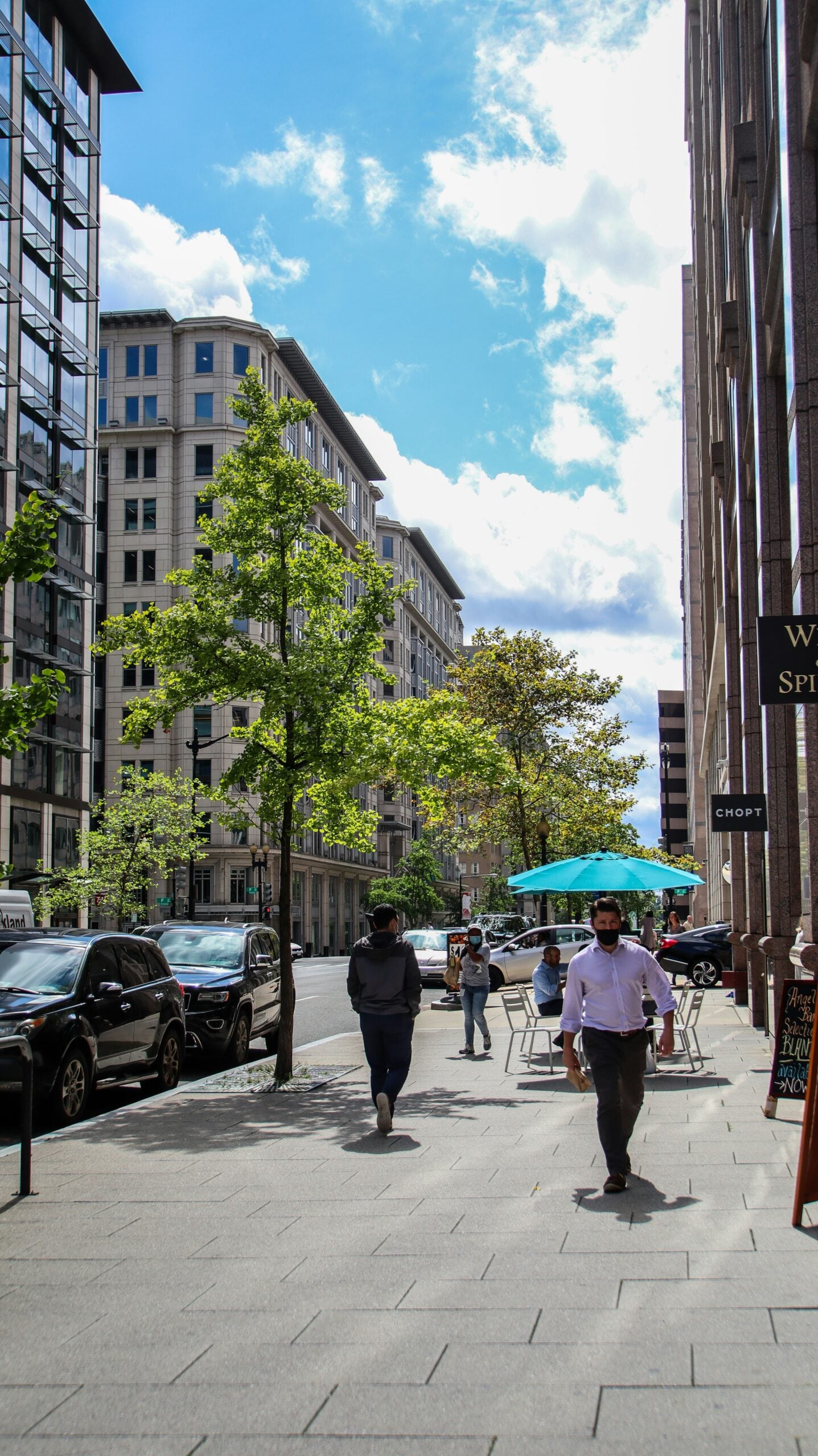 A city street scene with people walking on the sidewalk, parked cars, and outdoor tables with a turquoise umbrella. Tall buildings and trees line the street under a partly cloudy sky.