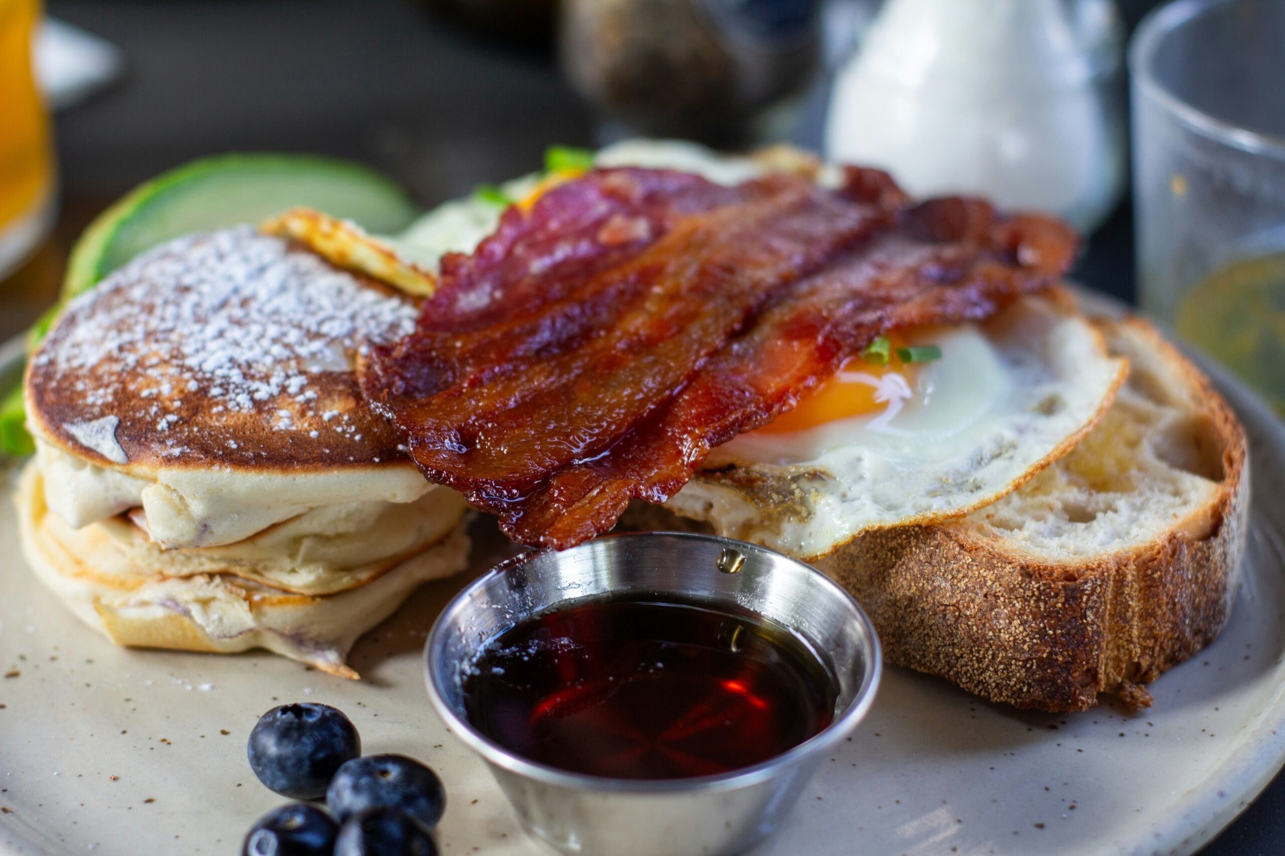 A breakfast plate with pancakes topped with powdered sugar, crispy bacon, a sunny-side-up egg on toast, a small metal cup of syrup, and fresh blueberries on the side.