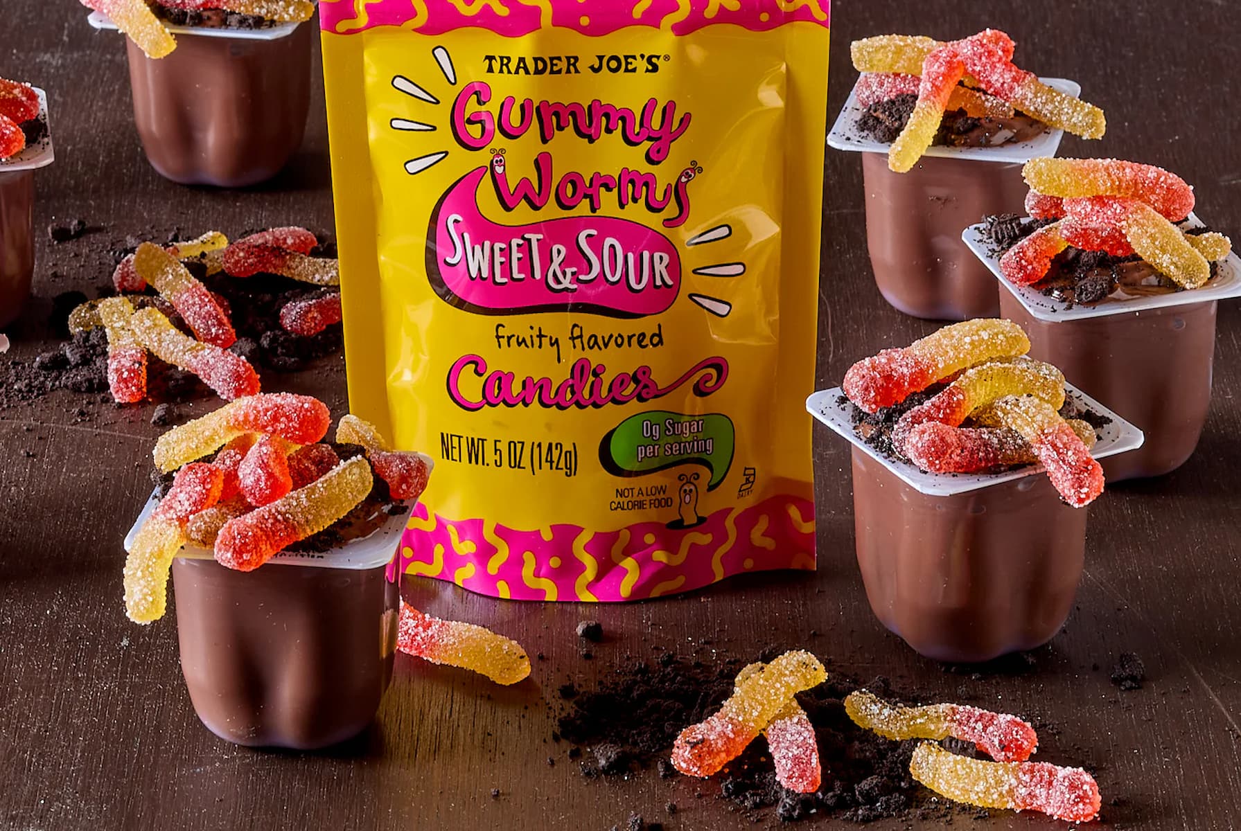 A bag of Trader Joe’s Gummy Worms is surrounded by chocolate pudding cups topped with crushed cookies and gummy worms, with extra gummy worms and cookie crumbs scattered on a wooden surface.