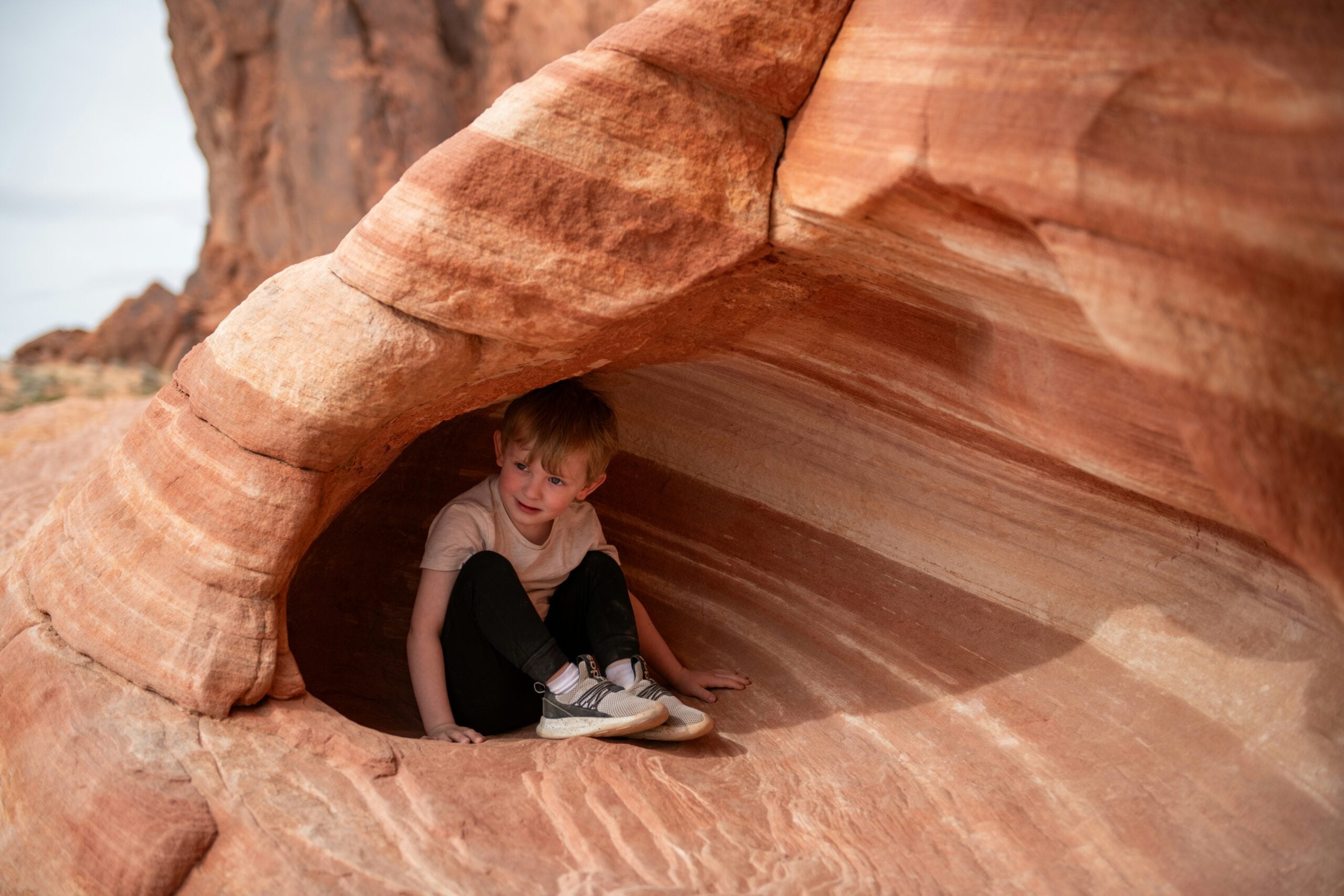 A young boy sits inside a small, naturally formed cave in red and tan striped sandstone, with sunlight illuminating the rock and the boy looking outward.