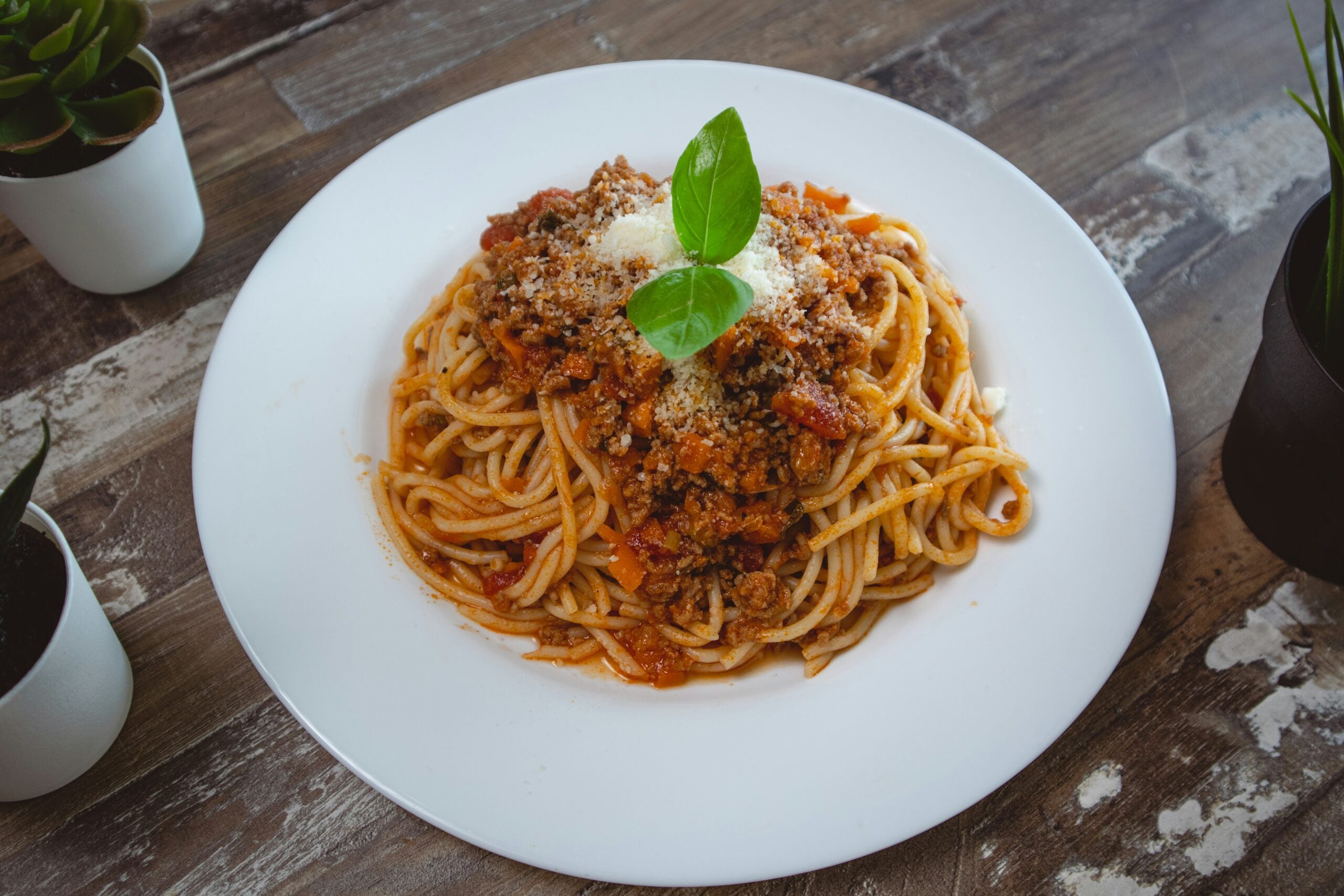 A plate of spaghetti topped with meat sauce, grated cheese, and fresh basil sits on a rustic wooden table, surrounded by small potted plants.
