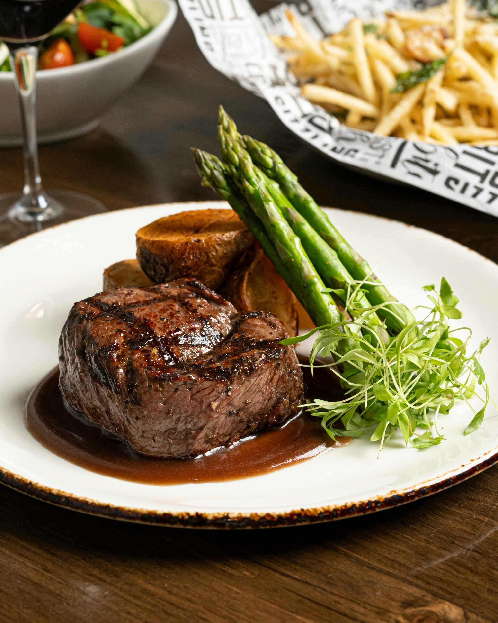 A plated meal featuring a grilled steak with brown sauce, roasted potatoes, fresh asparagus, and microgreens, with a side of fries and a bowl of salad in the background.