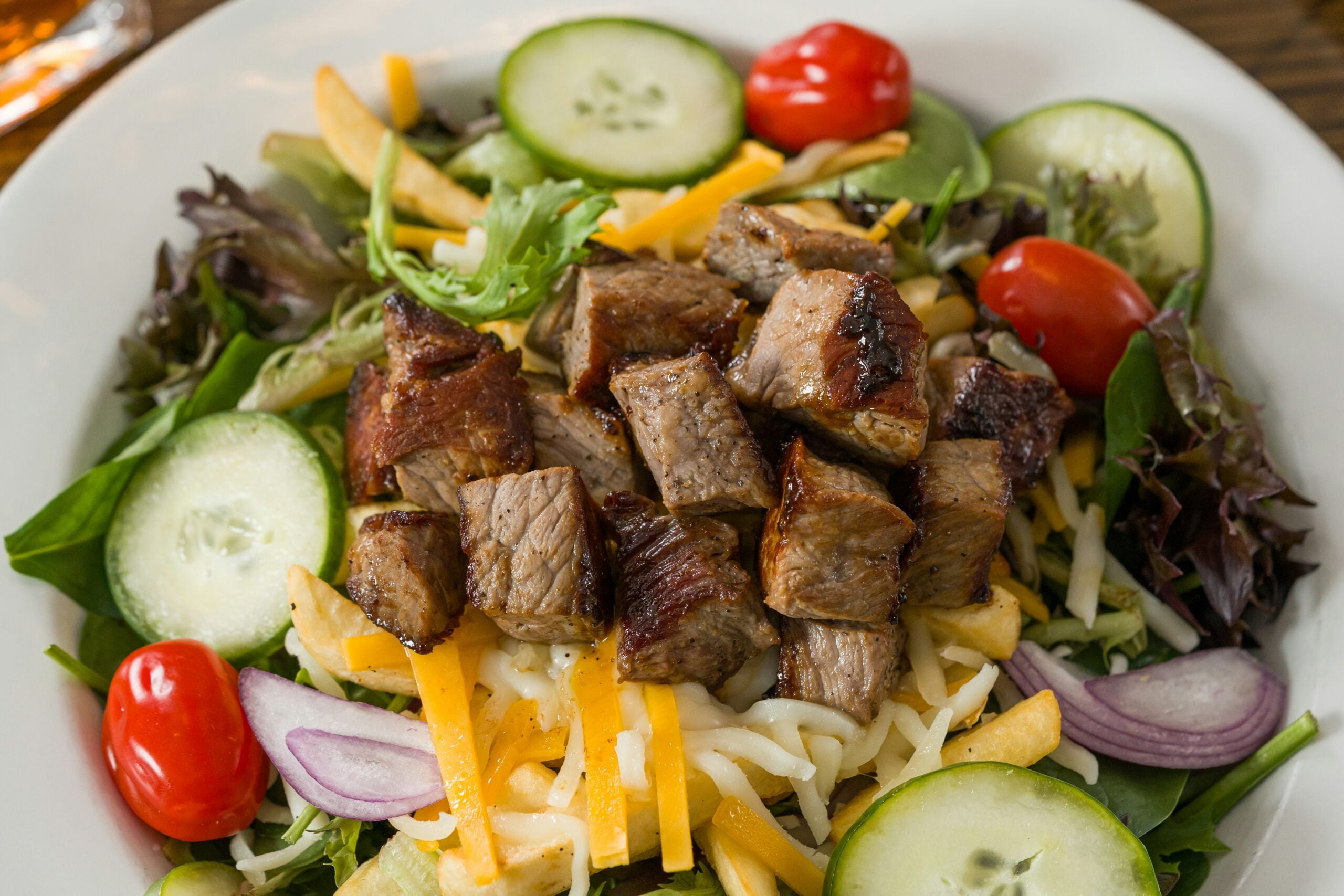A salad topped with grilled steak cubes, shredded cheese, cucumber slices, grape tomatoes, red onion, and mixed greens on a white plate.