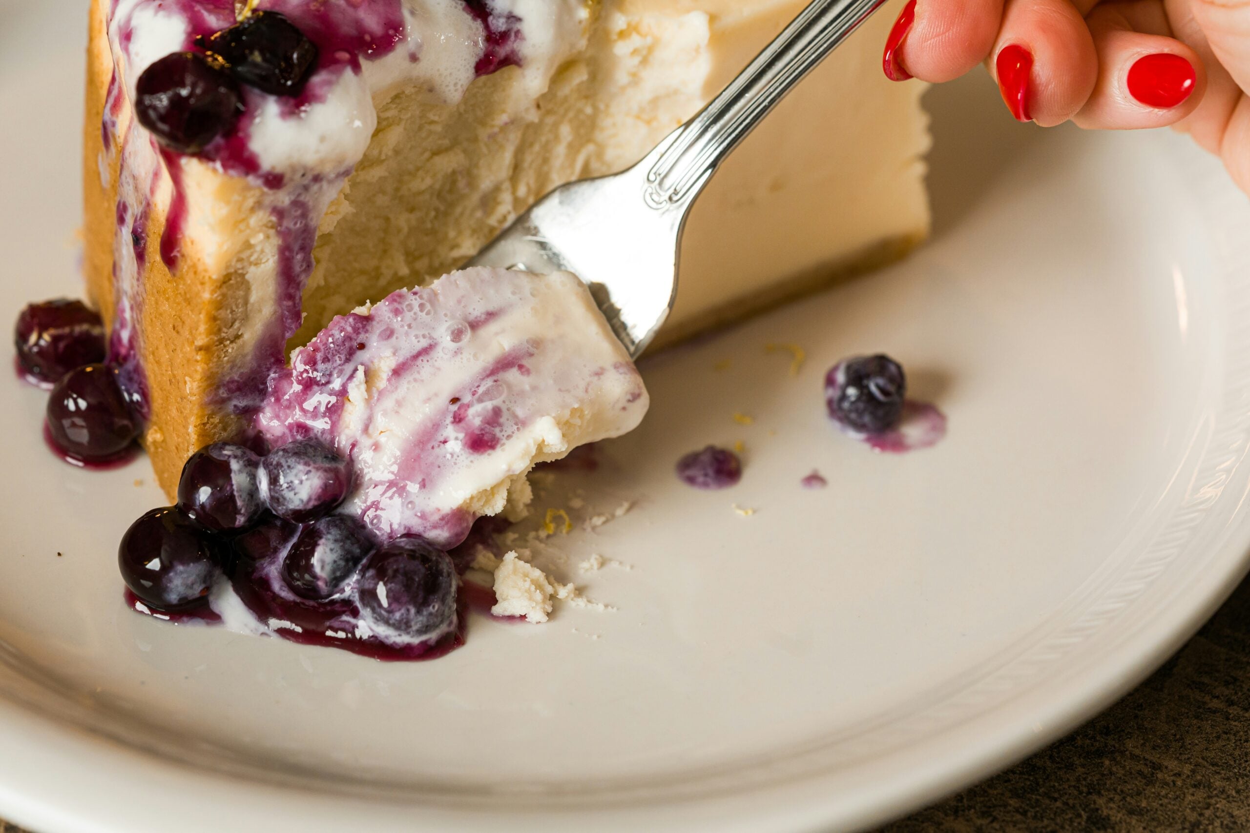 A hand with red nail polish holds a fork taking a bite of creamy cheesecake topped with blueberries and blueberry sauce, served on a white plate.