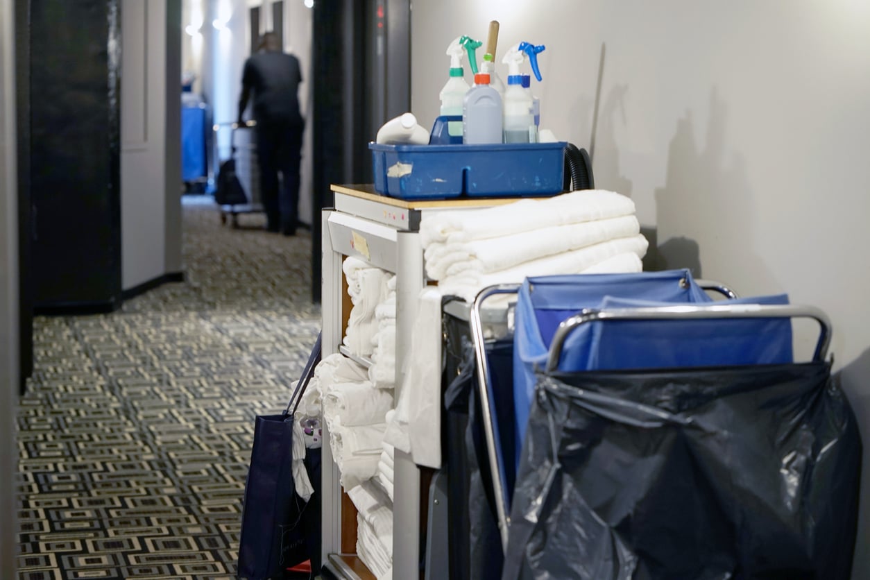 A hotel hallway with a housekeeping cart stacked with towels, cleaning bottles, and a large laundry bag—reminding guests to practice good hotel room etiquette. A person is visible in the background walking away down the corridor.