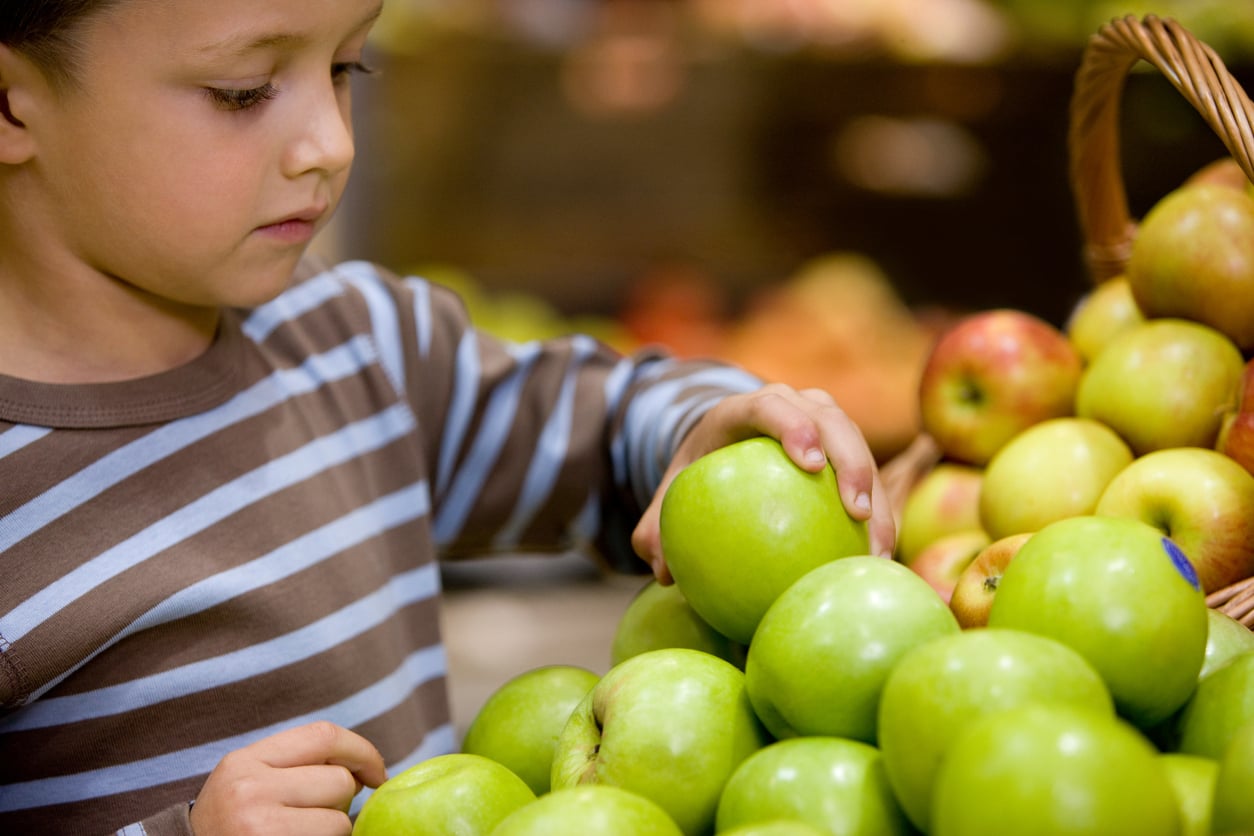 A child wearing a brown and white striped shirt selects a green apple from a pile of apples at a market or grocery store.
