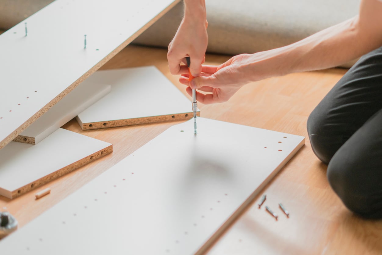 A person kneels on the floor, using a screwdriver to assemble white particleboard furniture panels, with screws and additional panels scattered nearby.