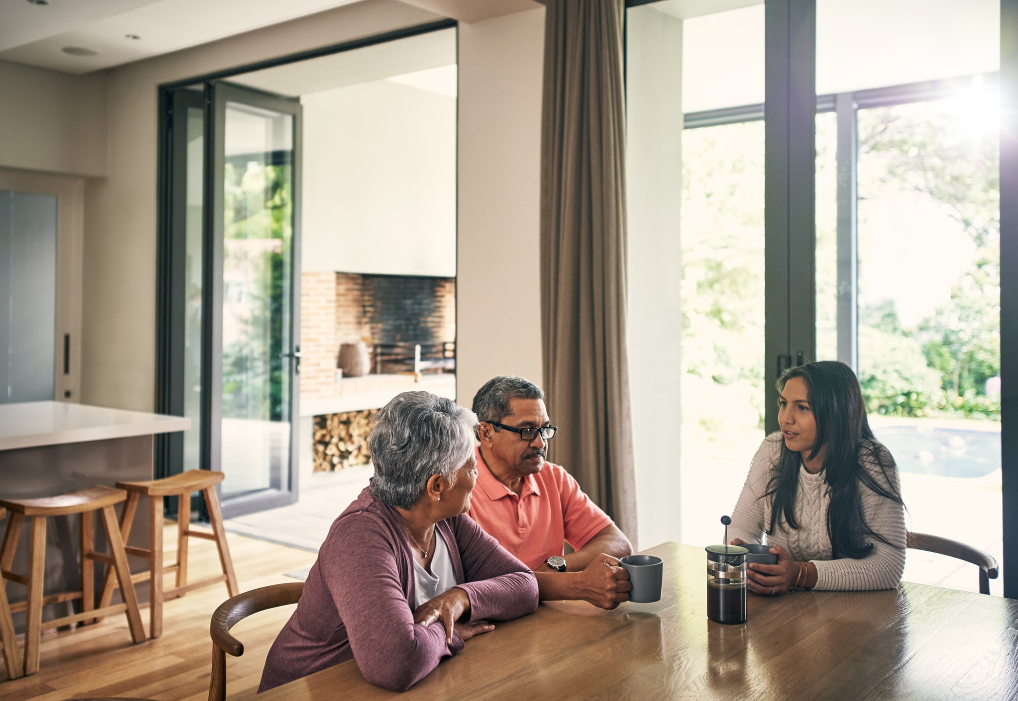 Three people sit at a wooden table in a bright, modern kitchen, engaged in conversation about how to support adult children financially. Two older adults and a younger woman enjoy coffee as sunlight streams through large glass doors.