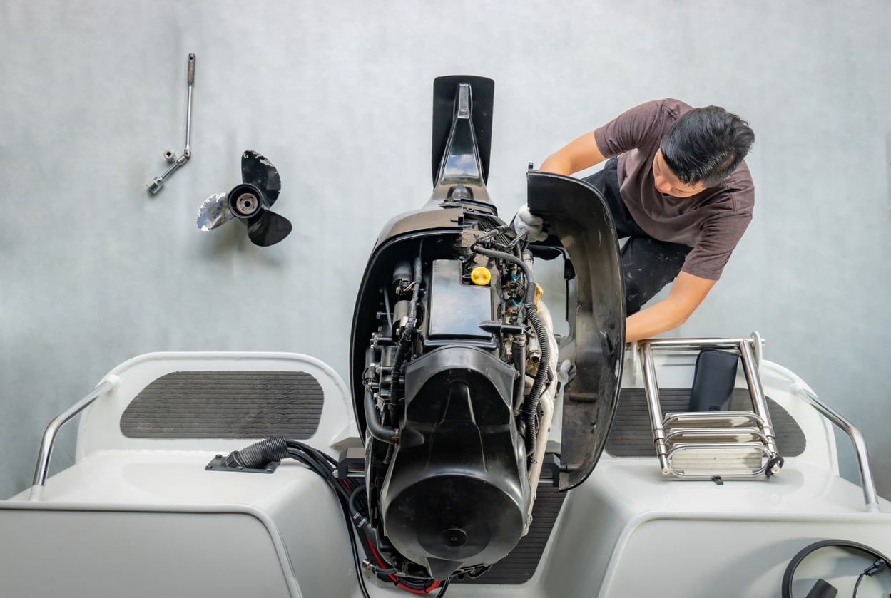 A person is repairing a boat engine from above, with tools and a propeller placed nearby on a white surface. The engine cover is removed, exposing internal components.