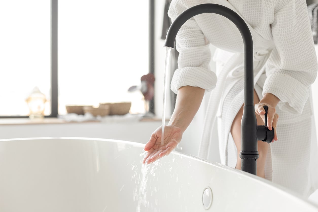 A person in a white bathrobe demonstrates hotel room etiquette by testing the water temperature with their hand under a black faucet, filling a white bathtub in a bright, modern bathroom.