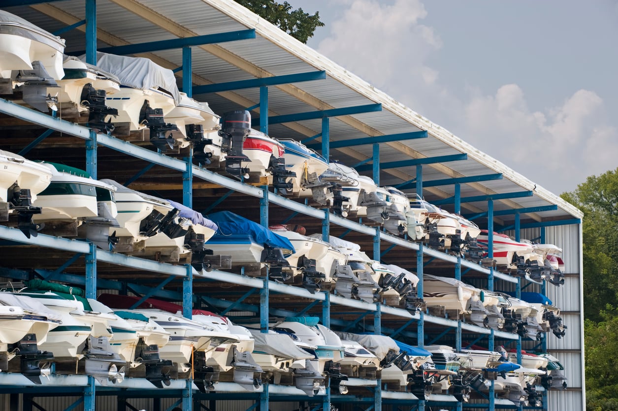 A large boat storage facility with multiple levels of motorboats stacked in metal racks, each boat covered or uncovered, beside a tree and under a partly cloudy sky.