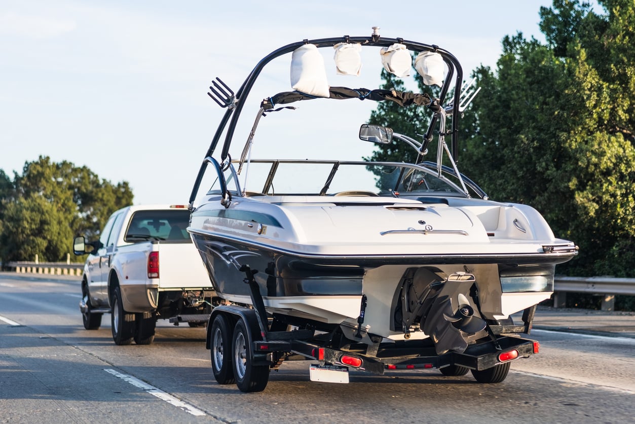 A white pickup truck is towing a motorboat on a trailer down a highway. The boat has a black and white hull and is equipped with speakers and a wakeboard tower. Trees and a guardrail line the road in the background.
