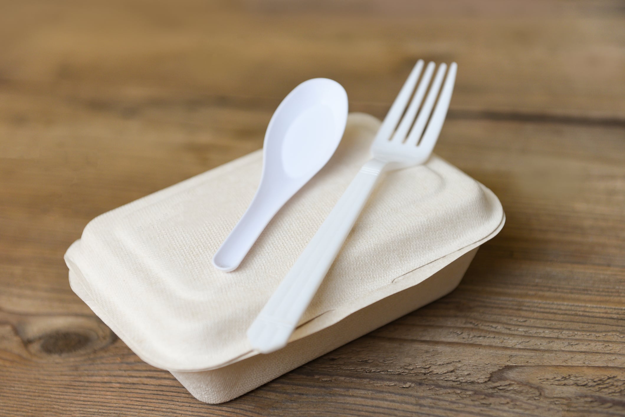 A closed, beige takeout food container sits on a wooden table, with a white plastic fork and spoon placed on top of the lid.