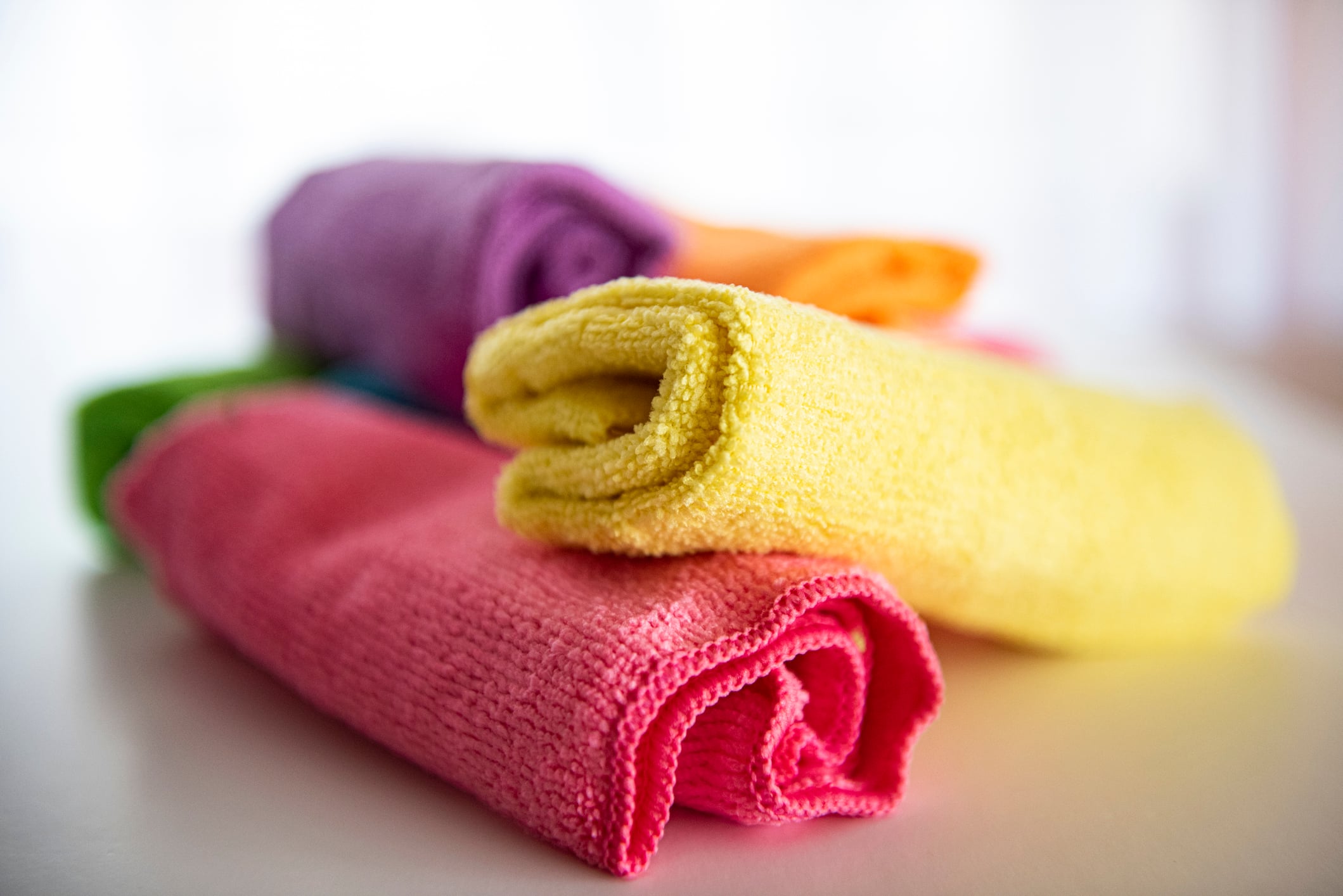 Several rolled microfiber cloths in bright colors, including pink, yellow, orange, green, and purple, are arranged on a white surface with a soft, blurred background.