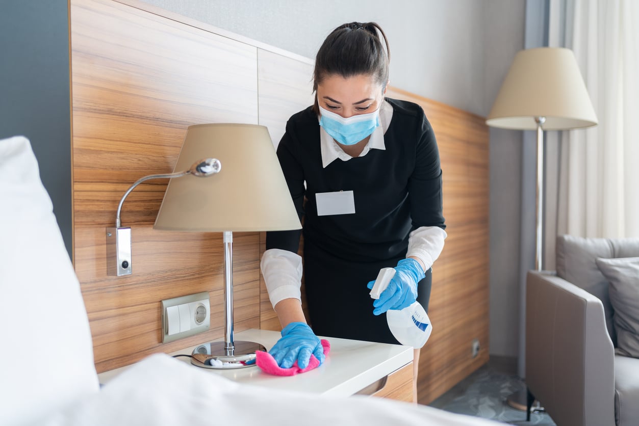 A woman wearing a face mask, gloves, and a uniform demonstrates hotel room etiquette as she cleans a bedside table with a spray bottle and pink cloth. A lamp and bed are visible in the modern, tidy room.