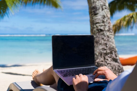 A person lounging on a chair under a palm tree works on a laptop, facing a tropical beach with clear blue water and a bright sky in the background.