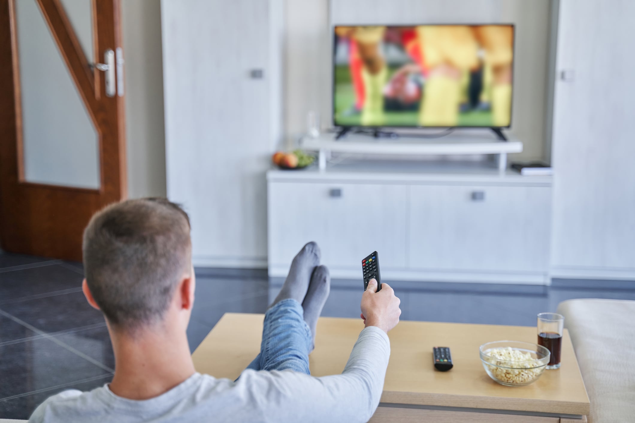 A person sits on a couch with feet up on a coffee table, holding a remote and watching TV. A bowl of popcorn and a glass of drink are on the table, and a sports game is on the television in a bright living room.