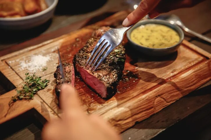 A person cuts into a juicy, medium-rare steak on a wooden board, garnished with herbs, with a small bowl of yellow sauce and a sprinkle of sea salt on the side.