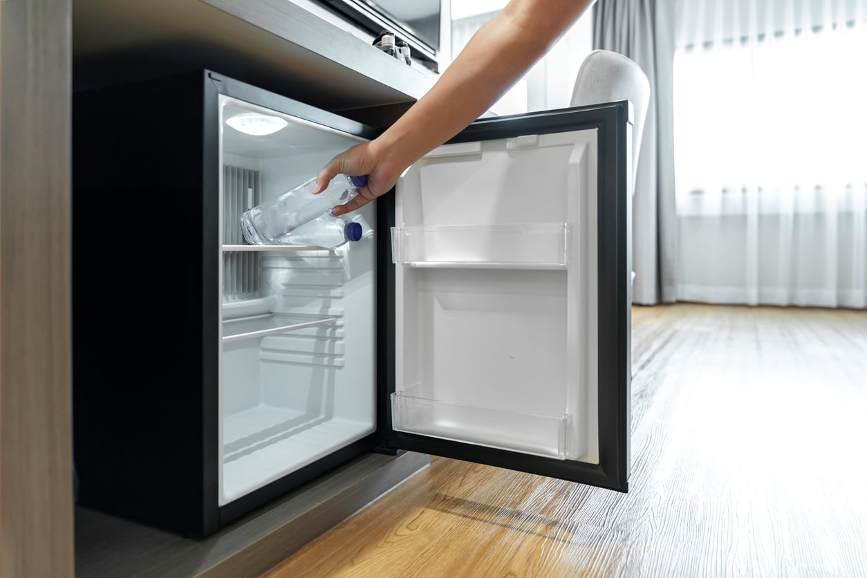 A person places a clear plastic water bottle inside an open, empty mini fridge situated under a desk in a bright room with wooden floors and large windows.