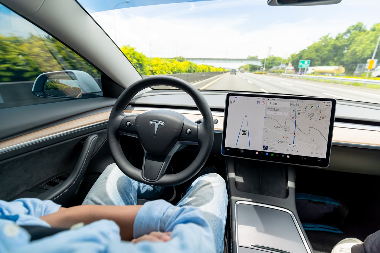 Person sitting in the driver’s seat of a Tesla with hands off the wheel, using autopilot mode; a large touchscreen display shows navigation and road details as the car travels on a highway.