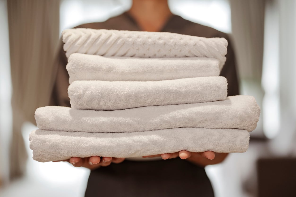 A person in a uniform holds a neatly stacked pile of clean, white towels with both hands. The softly blurred background suggests an indoor setting, emphasizing hotel room etiquette in a serene hotel or spa environment.
