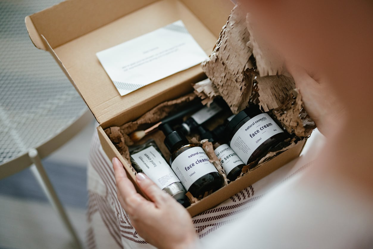 A person holds an open cardboard box filled with skincare products, including bottles and jars labeled "face cream," nestled in shredded brown packing paper. A folded note is also included in the box.