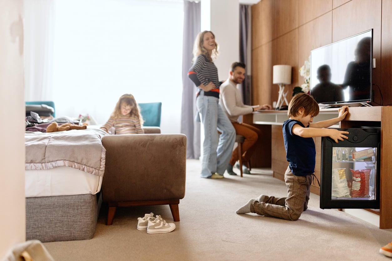 A young boy kneels to open a mini fridge in a hotel room, practicing good hotel room etiquette, while a girl sits on the bed and two adults smile in the cozy, bright space.