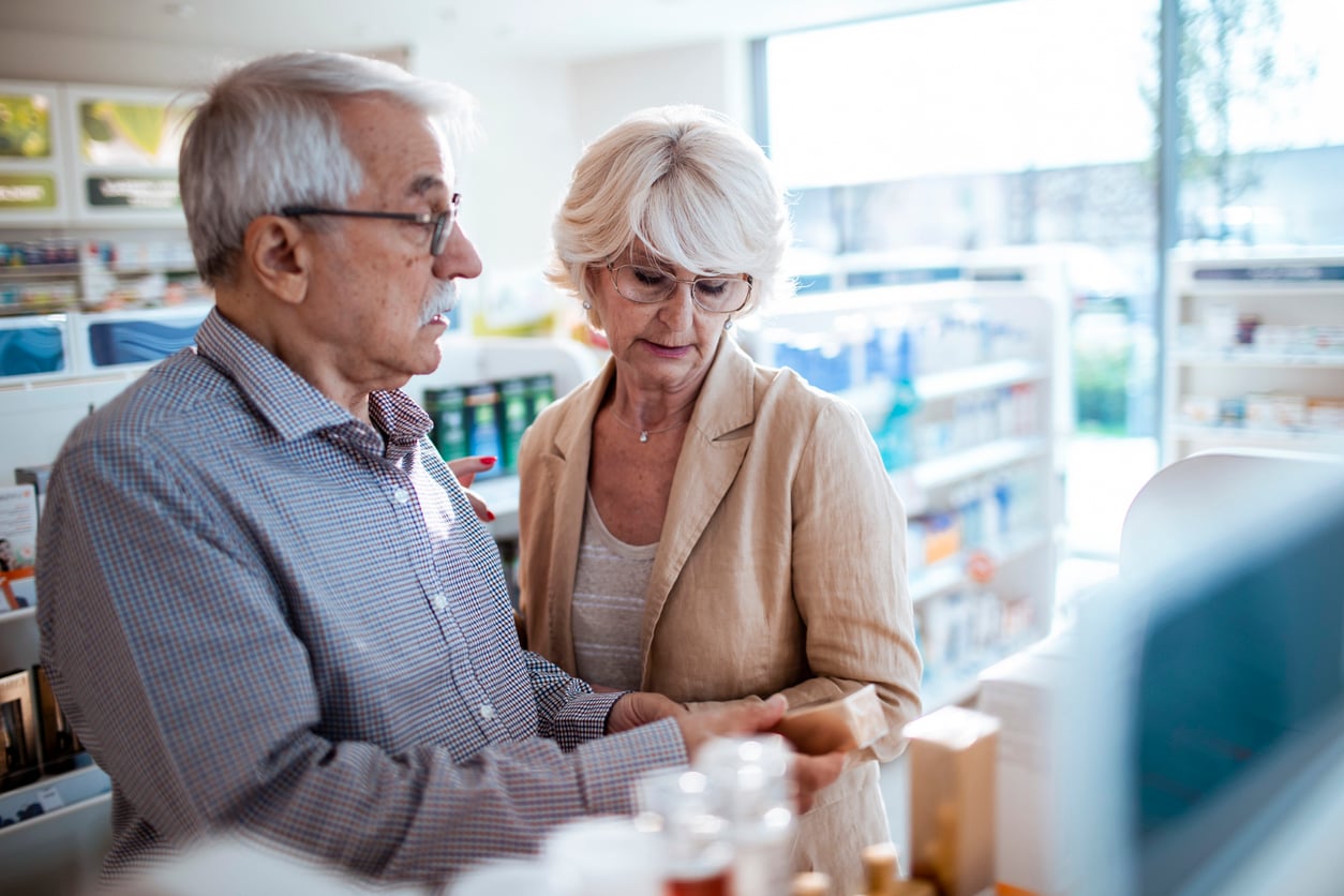 An elderly couple stands together in a pharmacy, closely examining a product while appearing to discuss it. Shelves with medications and health products are visible in the background.