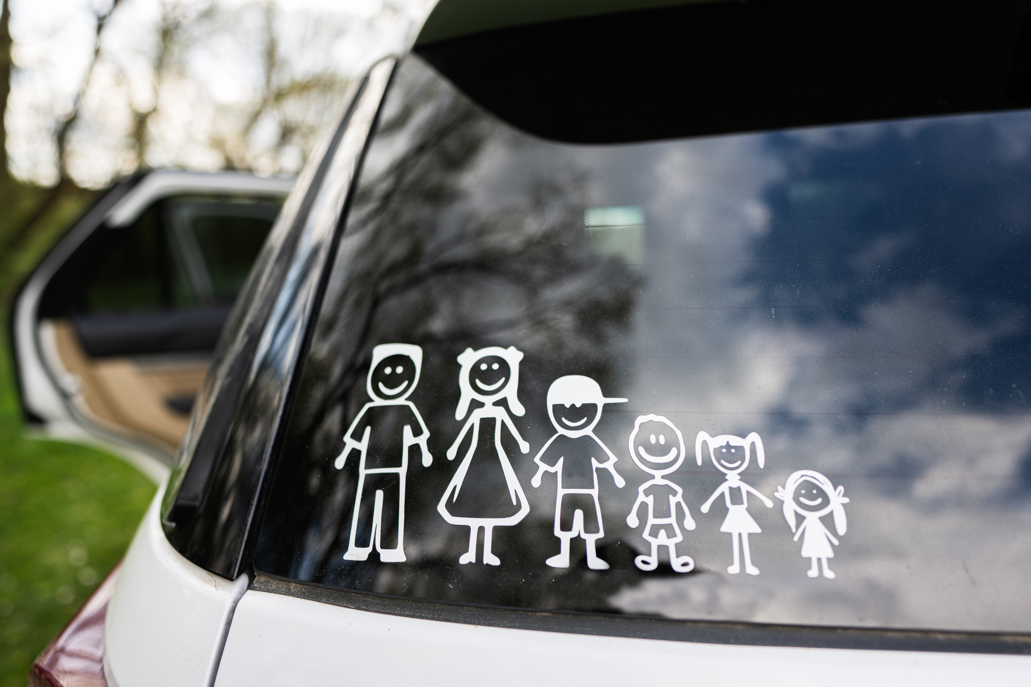 A car window displays white stick figure decals representing a family of five: two adults, two children, and a toddler, with trees and an open car door visible in the background.