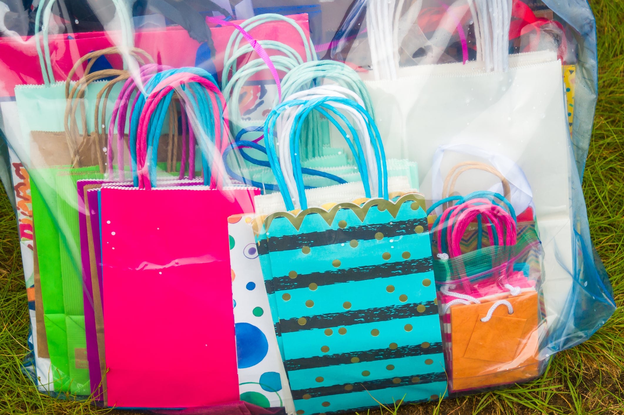 A variety of colorful gift bags with different patterns and designs are stacked together inside a transparent plastic container placed on green grass.