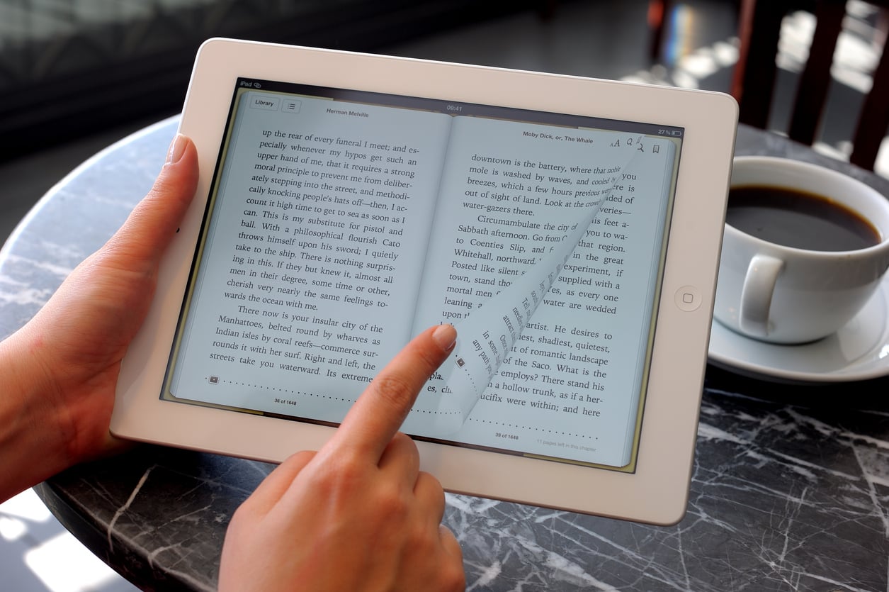 A person uses a tablet to read an e-book, swiping to turn the page, while sitting at a marble table with a cup of black coffee nearby.