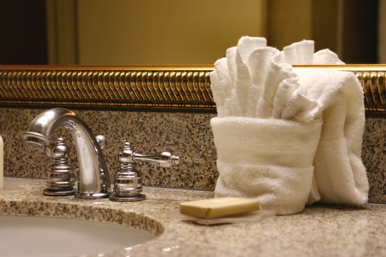 A bathroom sink with a silver faucet, a bar of soap on the granite countertop, and white towels neatly folded in a decorative arrangement next to a large framed mirror highlight proper hotel room etiquette.