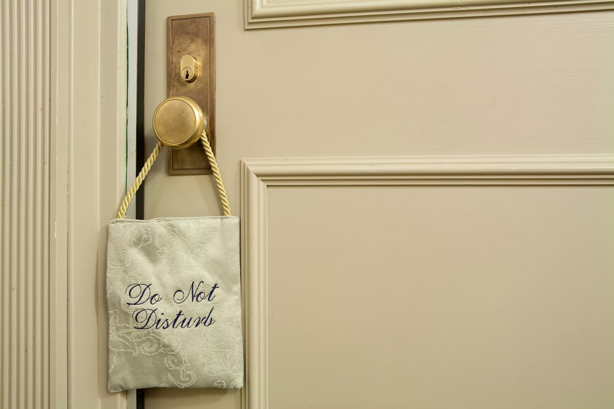 A gold door handle on a cream-colored door with a fabric "Do Not Disturb" sign in blue script perfectly illustrates hotel room etiquette.