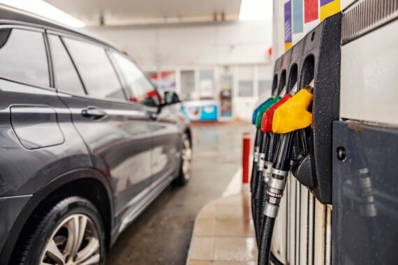 A close-up of fuel pumps with different colored handles at a gas station, next to a dark gray car parked beside the pump on a rainy day. The gas station building is visible in the background.