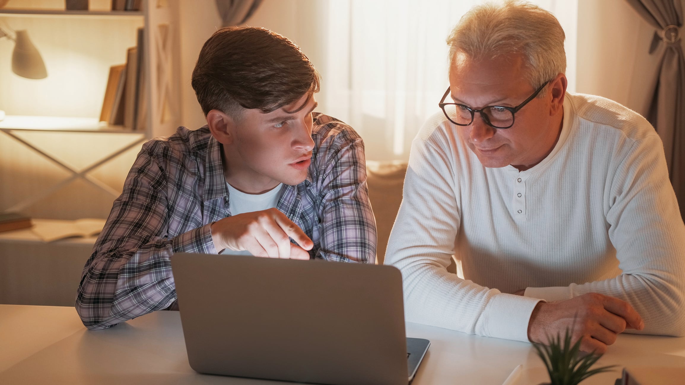 A young man and an older man sit side by side at a table, looking at a laptop screen. The young man is pointing at the screen while explaining ways to support adult children financially, and the older man listens attentively.