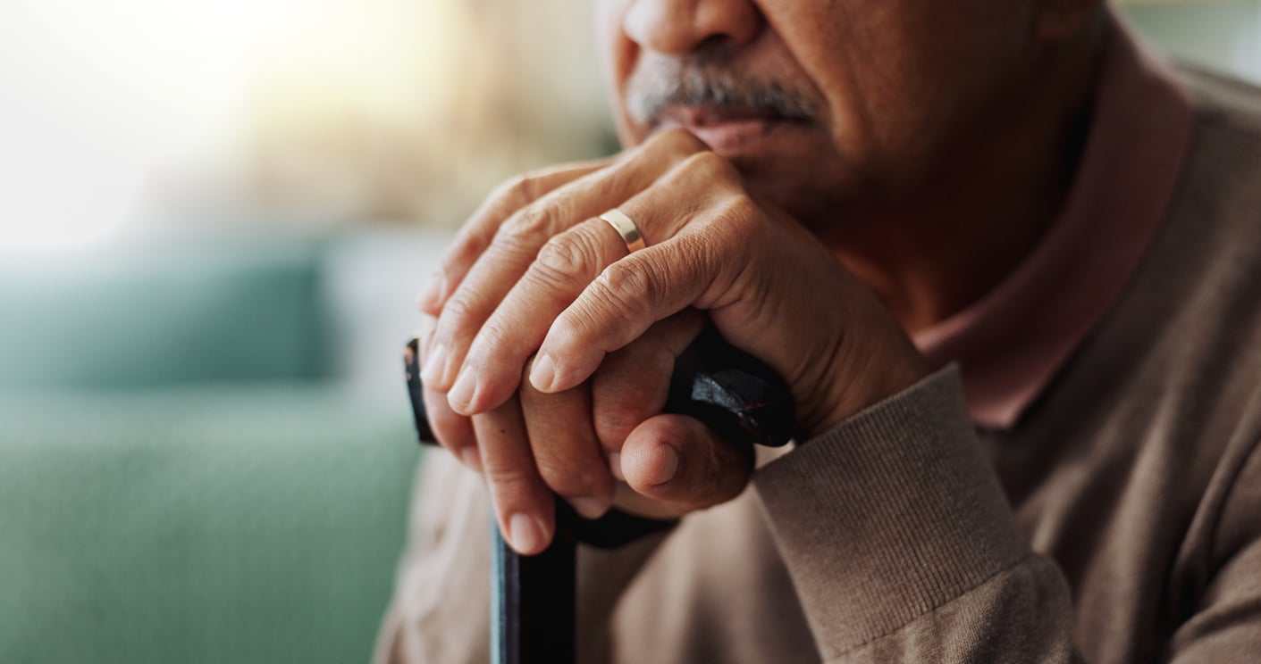 Close-up of an elderly man's hands resting on top of a cane, wearing a wedding ring, with his face partially visible and a soft, blurred background.
