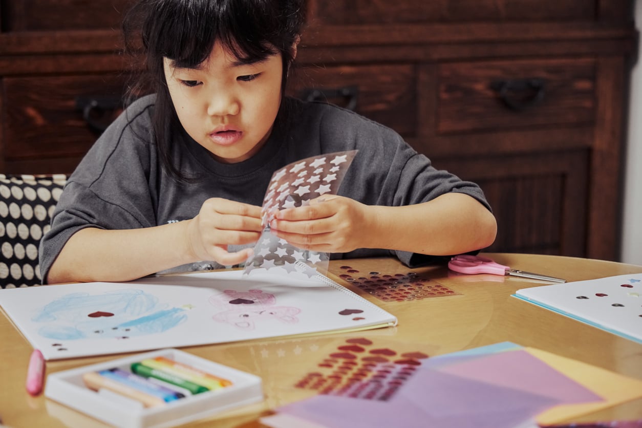 A young girl sits at a table, concentrating as she adds star stickers to a notebook filled with colorful drawings—even using some free things you can get at grocery stores, like sample crayons and fun envelopes scattered around her.
