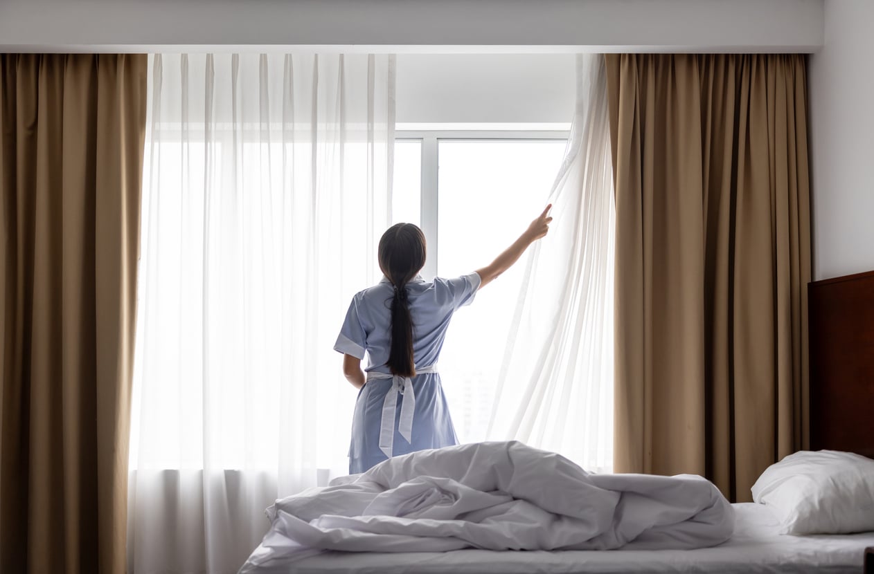 A woman in a uniform stands by a window, pulling aside white curtains in a hotel room with unmade bedding and brown drapes, letting in natural light—an example of proper hotel room etiquette.