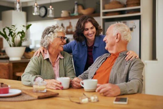A smiling older couple sits at a kitchen table with coffee, while a younger woman stands behind them, hugging them affectionately. All three people are happy, enjoying time together in a cozy home setting.